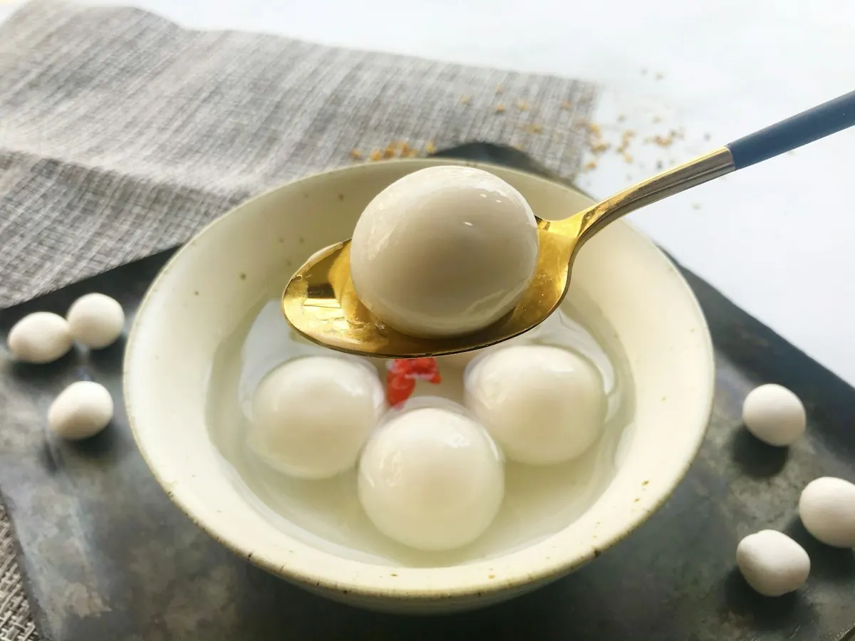 White glutinous rice balls in clear sweet soup, one lifted on a gold spoon